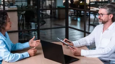 Dos personas conversan en una oficina frente a un ordenador portátil.