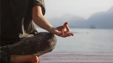 Persona meditando en posición de loto junto a un lago.