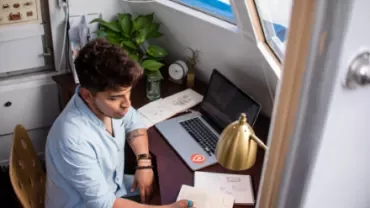 Persona escribiendo en un cuaderno junto a una computadora portátil.