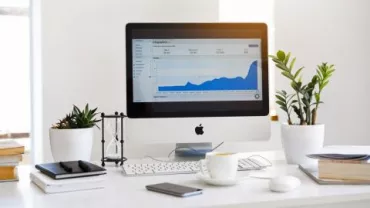 Escritorio con ordenador mostrando gráfico de datos, rodeado de plantas y libros.