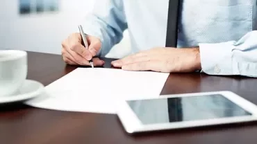 Persona escribiendo en un documento en una oficina con una taza de café y una tablet sobre la mesa.