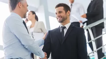 Dos personas dando la mano en una oficina mientras otras trabajan al fondo.
