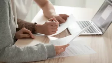 Dos personas revisando documentos frente a una laptop.