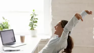 Persona estirándose en una oficina luminosa junto a una laptop.