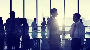 Varias personas de pie en una oficina, dos de ellas estrechan manos.