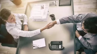 Dos personas estrechando manos en una mesa de oficina con documentos.