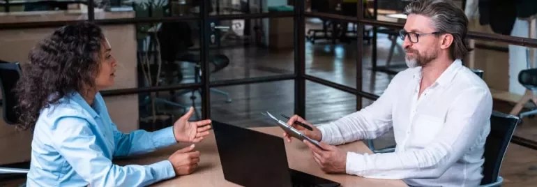 Dos personas conversan en una oficina frente a un ordenador portátil.