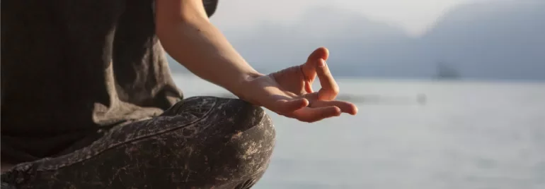 Persona meditando en posición de loto junto a un lago.