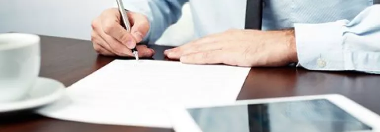 Persona escribiendo en un documento en una oficina con una taza de café y una tablet sobre la mesa.