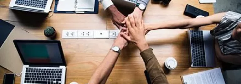 Manos unidas sobre una mesa con laptops.