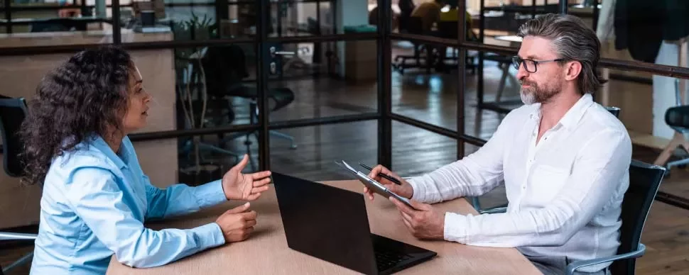 Dos personas conversan en una oficina frente a un ordenador portátil.