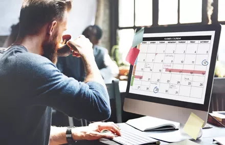 Persona trabajando en un escritorio con un calendario en la pantalla de la computadora.