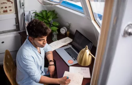 Persona escribiendo en un cuaderno junto a una computadora portátil.