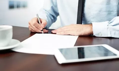 Persona escribiendo en un documento en una oficina con una taza de café y una tablet sobre la mesa.