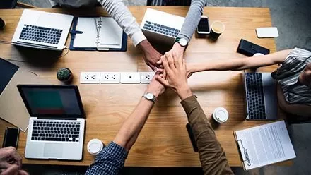 Manos unidas sobre una mesa con laptops.